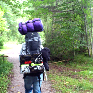 Foto einer Person mit vollbepackten Wanderrucksack, oben drauf eine Schlafsack und hinten dran etwas großes rechteckiges in einer Plastiktüte.