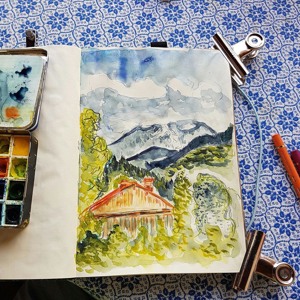 Buntstift und Aquarell auf Notizbuchpapier.Foto: Skizzenbuch mit bunter alpiner Zeichnung. An den Ecken Metallklammern und links ein Miniaquarellkasten.