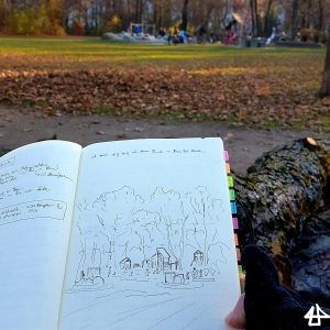 Foto: geöffnetes Skizzenbuch mit Füller-Zeichnung von Spielplatz unter großen Bäumen, im Hintergrund dieser Spielplatz.