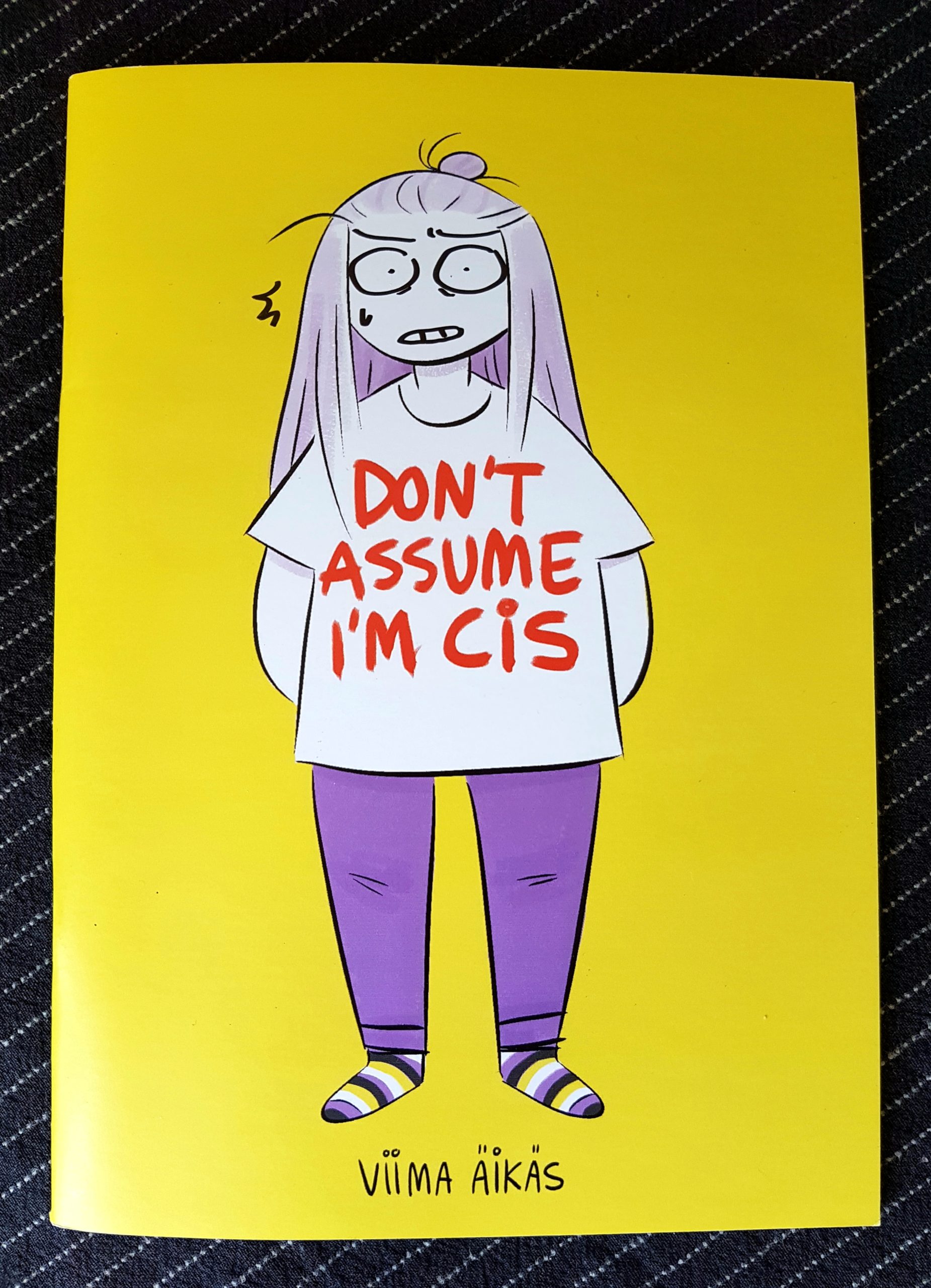 Buchdeckel von »Don't assume I'm cis« von Viima Äikäs. Eine Comiczeichnung einer weißen Person mit langen Haaren, xier trägt den Buchtitel rot gedruckt auf dem T-Shirt, deren Hose ist lila, der Hintergrund gelb und die Socken geringelt in den Farben der non-binary Flagge: gelb, weiß, lila, schwarz.