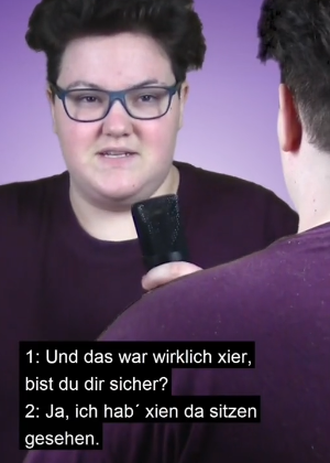 Screenshot aus dem Tiktok Video, der gesprochene Text ist eingeblendet: Person mit rundem Gesicht, dunklen Haaren und großer blauer Brille spricht in ein Mikrofon: Und das war wirklich xier, bist du dir sicher? Die Person davor wird nur von hinten gezeigt, trägt den gleichen dunklen Pullover und antwortet: Ja, ich hab xien da sitzen gesehen.