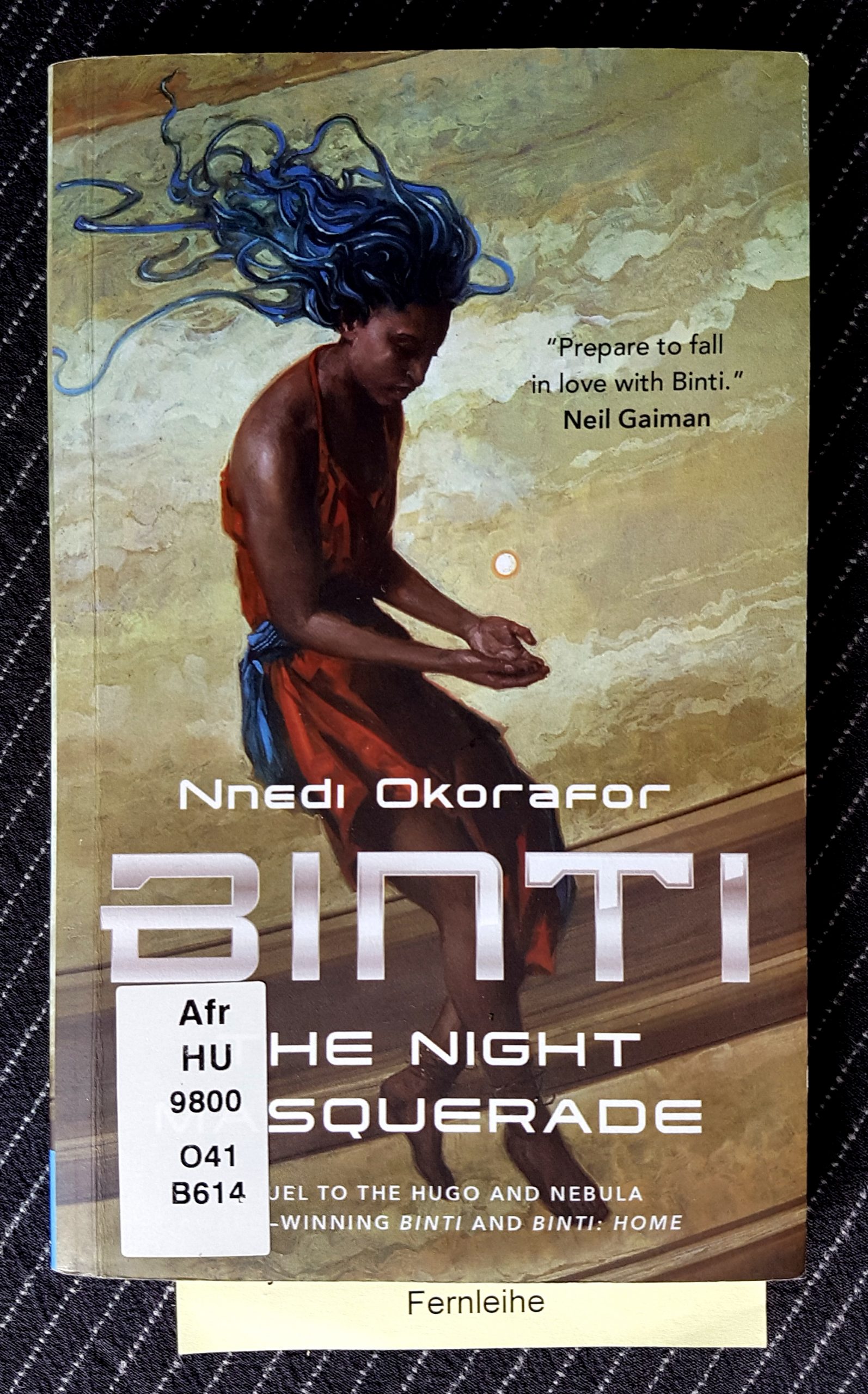 Buchdeckel von »Binti - The night Masquerade« von Nnedi Okorafor. Eine junge schwarze Person mit blauen tentakeligen Haaren und orangefarbenem Kleid sitzt auf einem hellen Steinsockel. Über den geöffneten Händen im Schoß schwebt eine weiße Kugel. Aus dem Buch guckt ein Zettel mit dem Wort Fernleihe.