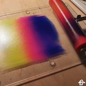 Kleine Glasplatte auf Holzoberfläche, darauf eine Linoldruckwalze und ein Farbverlauf von gelb nach lila als dünner Film auf der Glasplatte.