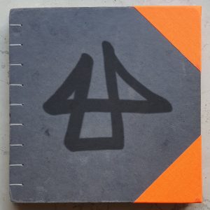 Foto vom Buchdeckel des Skizzenbuchs, am linken Rand die Fäden von der handgemachten Bindung, auf dem grauen Karton das künstlerische Logo, mit leuchtend orange-farbenen Eckenverstärkungen rechts.