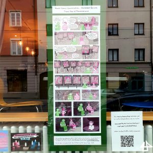 Foto eines langen Comics an einer Schaufensterscheibe. Daneben ein QR-Code und eine Postkarte mit hell-rosa, weißen und hellblauen erleuchteten Kerzen. Durch die Scheibe sind eine Heizung und Stühle sichtbar, in der Scheibe reflektiert außerdem die rote und die weiße Hausfassade von gegenüber.