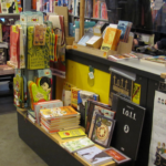 Minicomics-Box auf der Ladentheke im Modern Graphics Comic Laden voll kleiner und großer Tische mit Comics.