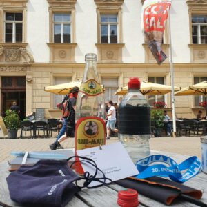 Foto aus der Erlanger Innenstadt, auf einem Kaffeehaustisch: Skizzenbuch, Wasserpinsel, Club Mate Flasche, Wasserflasche, Messeausweiss und FFP2 Maske. Dahinter ein sehr altes Haus mit Stuck und ein wehendes Banner des Comic Salons.