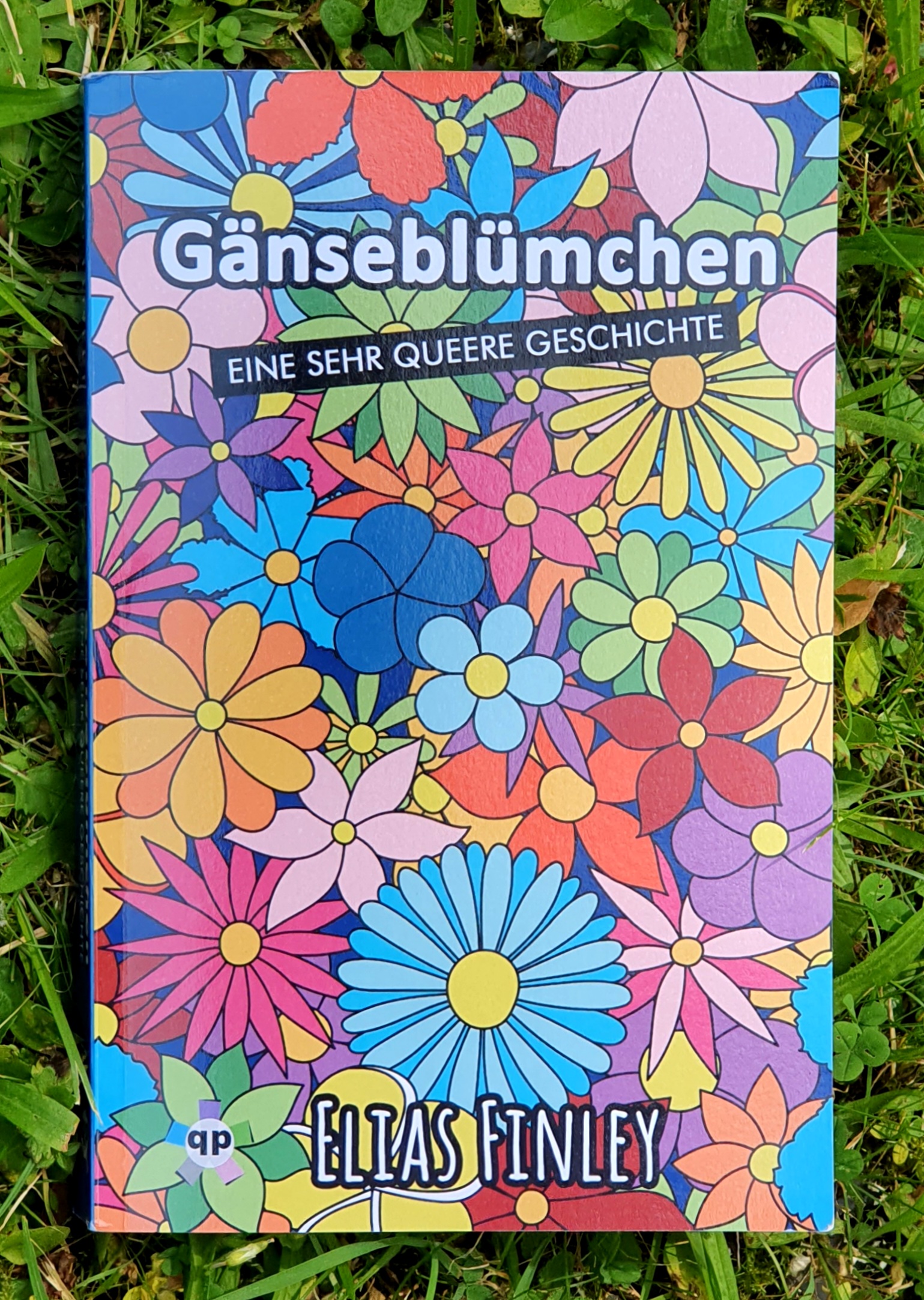 Foto eines Buches im Gras. Auf dem Umschlag ganz viele bunte gezeichnete Blumen. Text: Gänseblümchen - eine sehr queere Geschichte, qp Verlag, Elias Finley.