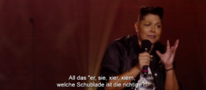 Sara Ramirez als Che Diaz in schwarzen Klamotten mit Mikrofon, darunter als Untertitel: All das 'er, sie, xier, xiem', welche Schublade ist die richtige?