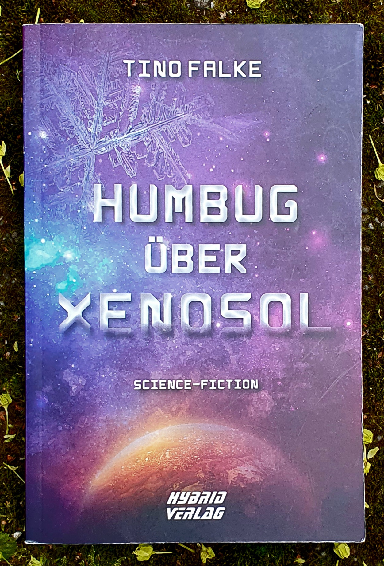 Foto des Buch-Deckels: Ein im Weltall rot-orange glühender Planet im Weltraum wird wie durch eine Scheibe beobachtet auf der Eiskristalle wachsen. Text: Tino Falke, Humbug über Xenosol, Science-Fiction, Hybrid Verlag.