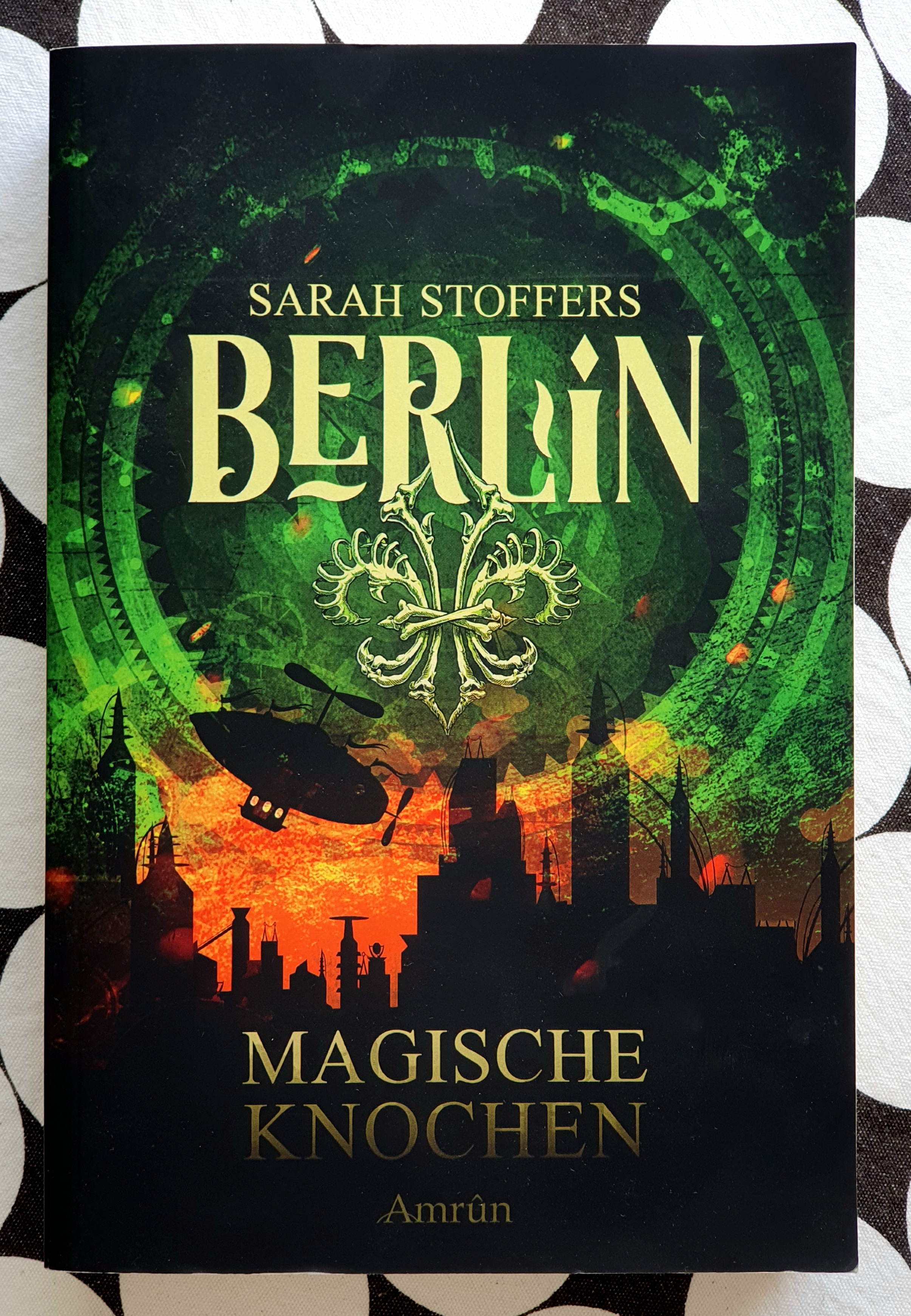 Foto des Einbandes von "Berlin - Magische Knochen. Unten eine schwarze Skyline einer Stadt mit vielen Türmen, darauf der Titel "Magische Knochen" und der Verlag Amrun. Das Wort Berlin und der Name Sarah Stoffers stehen vor dem grün schimmernden Himmel an dem ein Zeppelin fliegt.