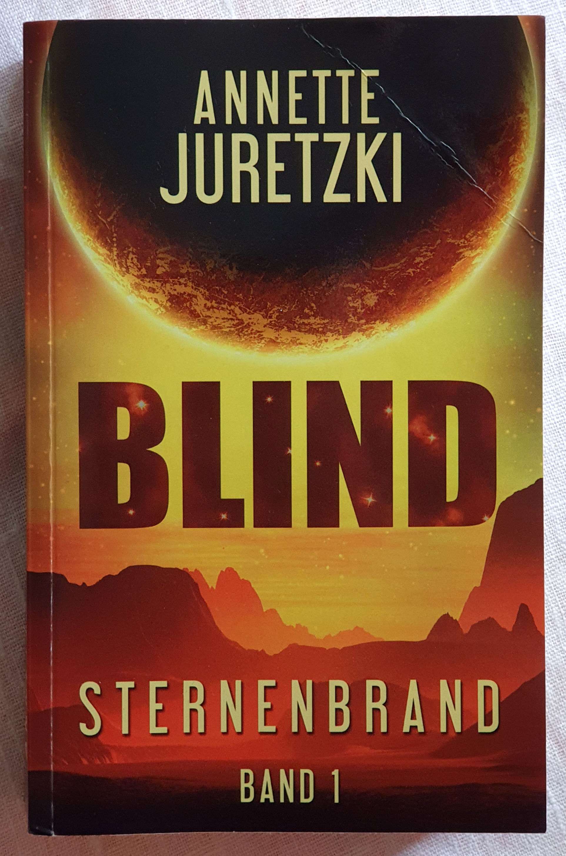 Deckblatt mit orangeroter Landschaft und einem großen dunklen Himmelskörper hinter dem die Sonne scheint. Text: Annette Juretzki, Blind, Sternenbrand, Band 1.
