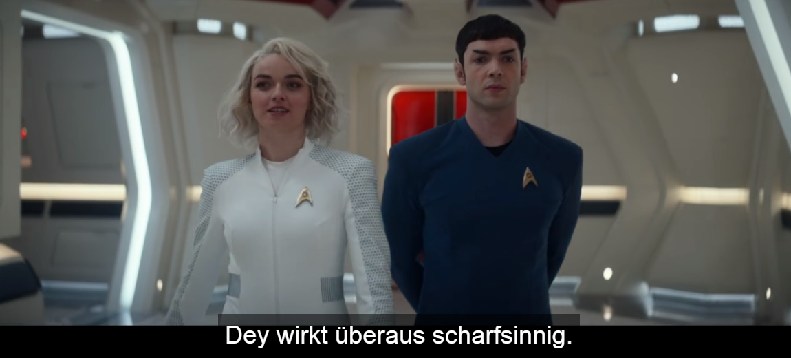 Screenshot aus der Serie. Mr. Spock in blauem Anzug läuft im Gang des Raumschiffes neben einer Person mit blonden halblangen Haaren. Untertitel: Dey wirkt überaus scharfsinnig.