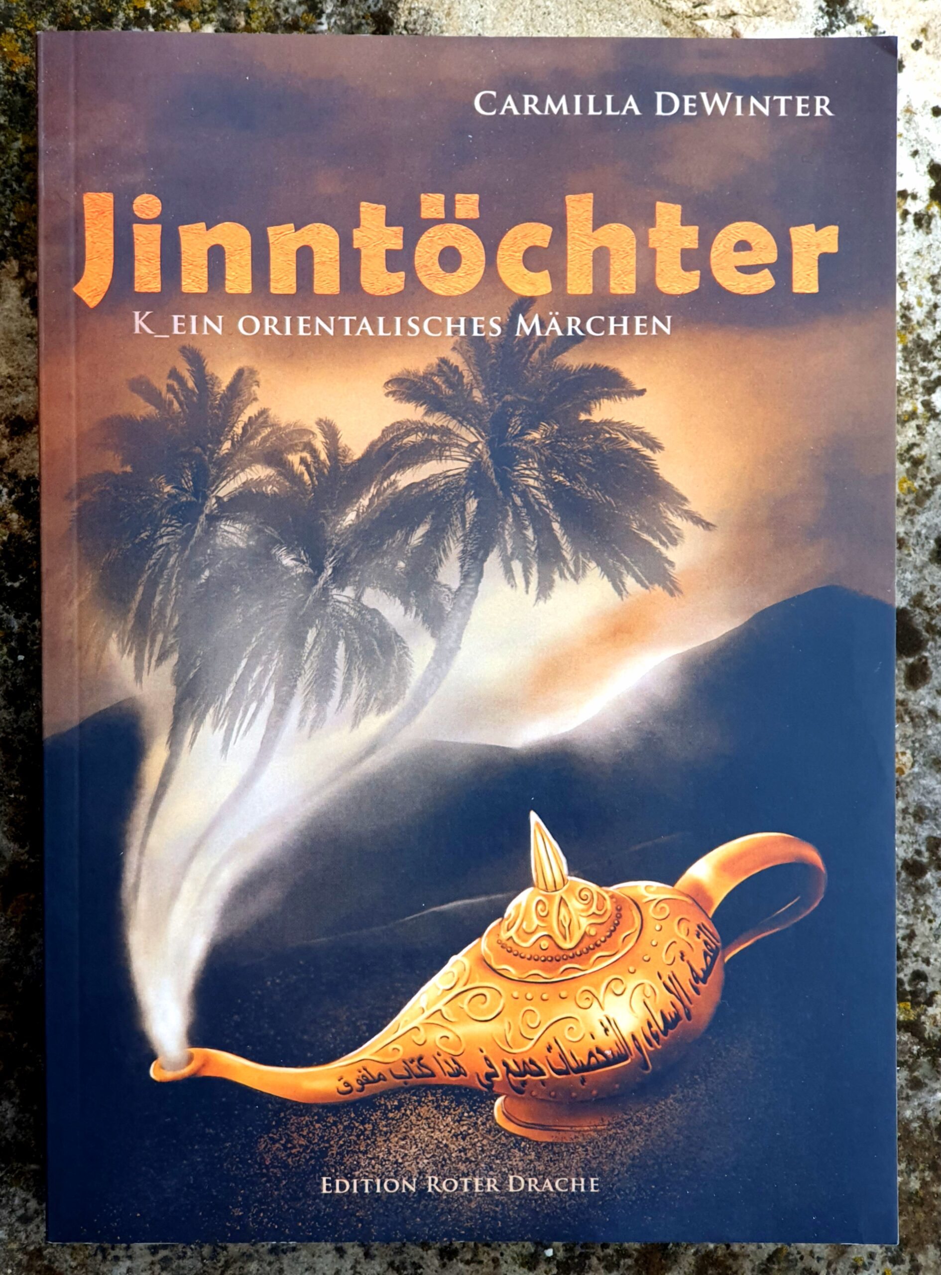 Buchdeckel des Romans von Carmilla DeWinter bei Edition Roter Drache: Jinntöchter - k_ein orientalisches Märchen. Der Titel sehr groß über einer Wüstenlandschaft mit dunklen Bergen. Im Sand eine goldene Öllampe aus der Rauch aus der Tülle steigt, im Rauch das vage Bild von drei Palmen.