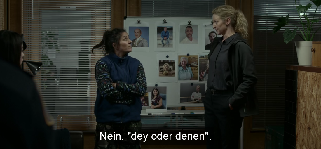 Screenshot aus der Serie: Dulcie mit blondem Pferdeschwanz und Eddie mit braunem Pferdeschwanz stehen sich vor einem Aufsteller mit Fotos gegenüber. Untertitel: Nein, dey oder denen.