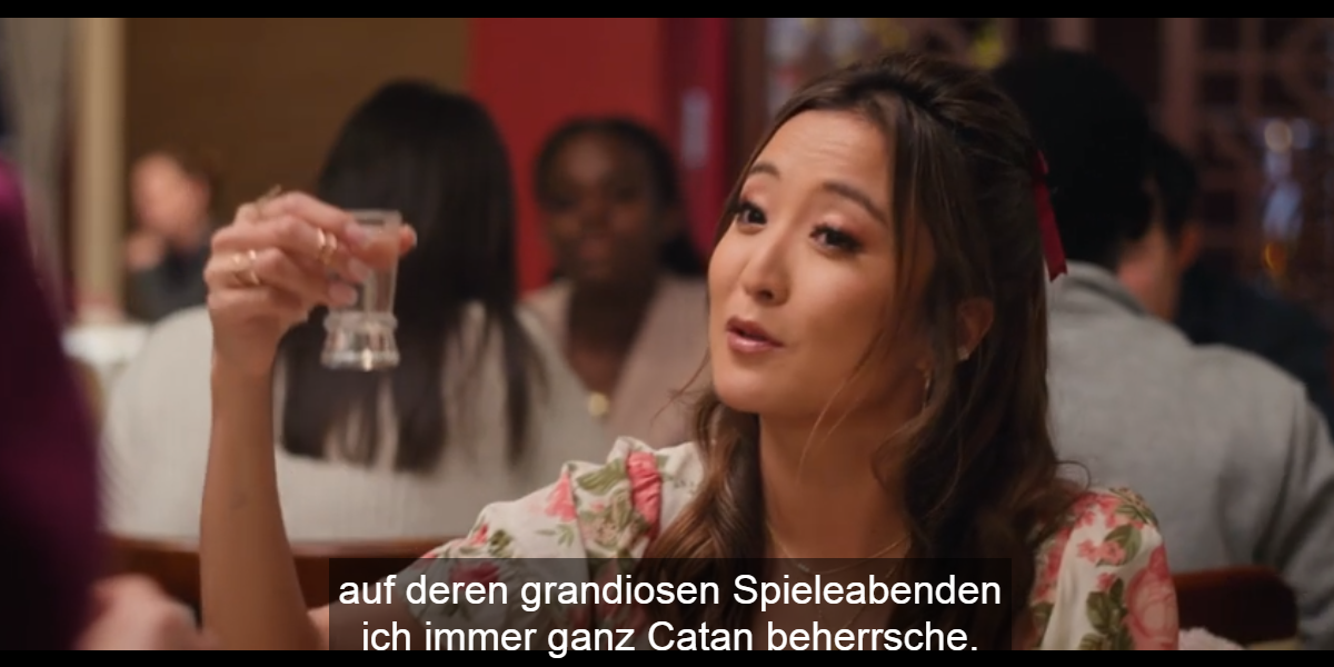 Screenshot aus dem Film. Ein Person mit langen braunen Haaren und geblühmter Bluse hebt ein Shotglas in einem Restaurant. Untertitel: auf deren grandiosen Spieleabenden ich immer ganz Catan beherrsche.