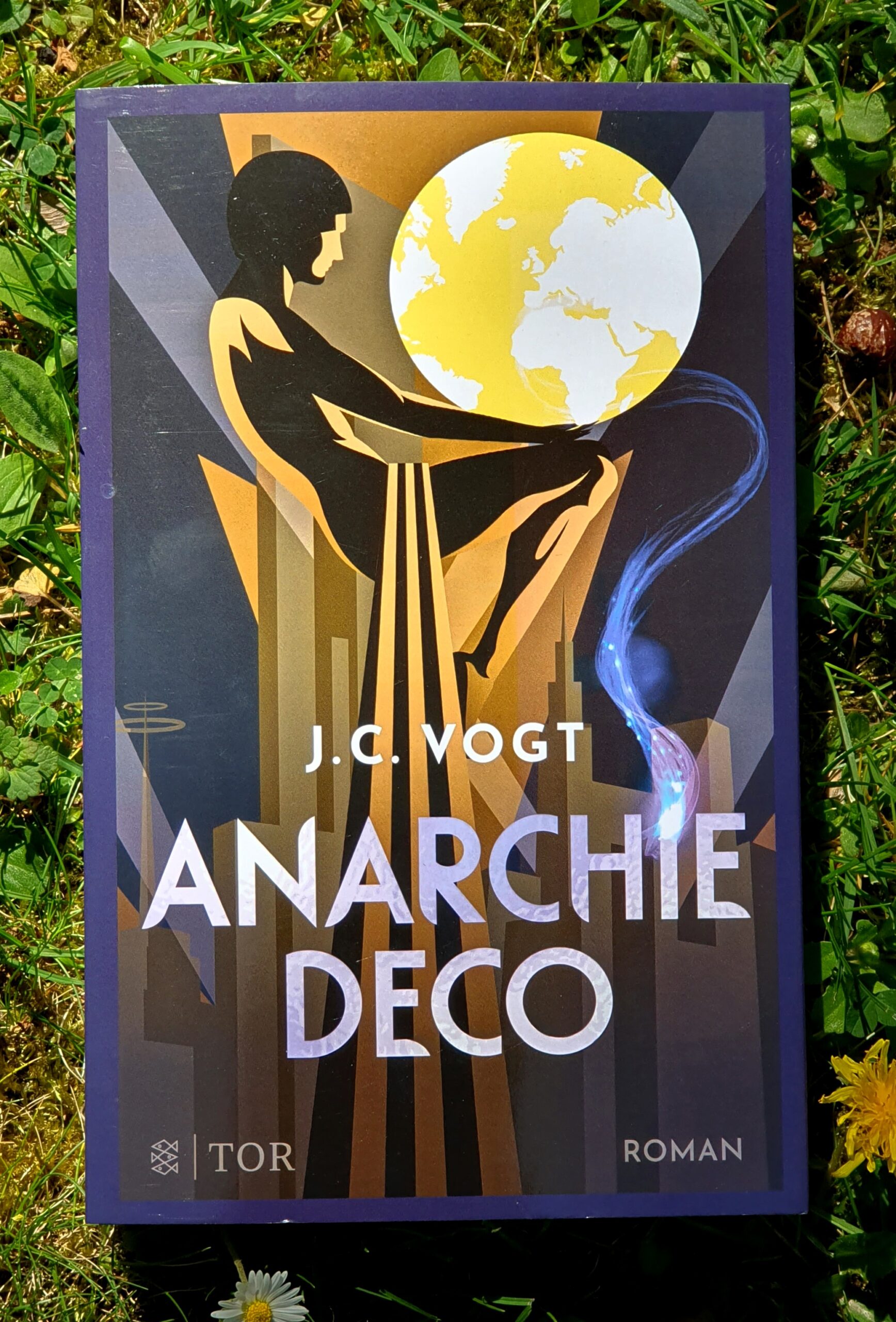 Buchcover im Gras fotografiert, "Anarchie Deco", Roman im TOR Verlag, von J.C. Vogt. Im Hintergrund der Schrift eine Art Statue, die sitzend den Planeten Erde hält. Es könnte auch eine Lampe sein.