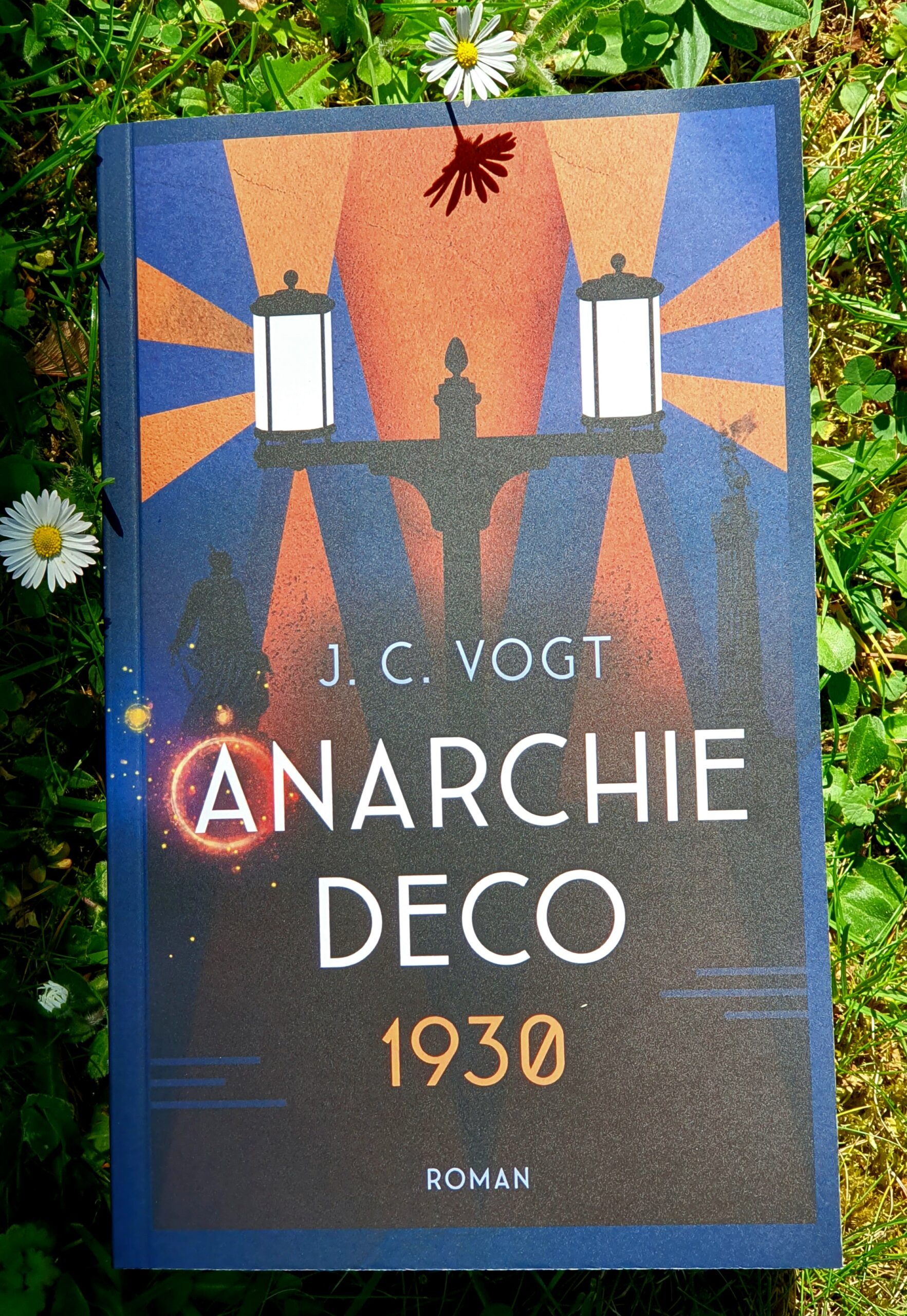 Buchcover im Gras fotografiert, "Anarchie Deco 1930", Roman von J.C. Vogt. Im Hintergrund der Schrift eine gusseiserne Laterne mit zwei hell erleuchteten Lampenschirmen, die orangenes Licht in die blaue Nacht werfen.