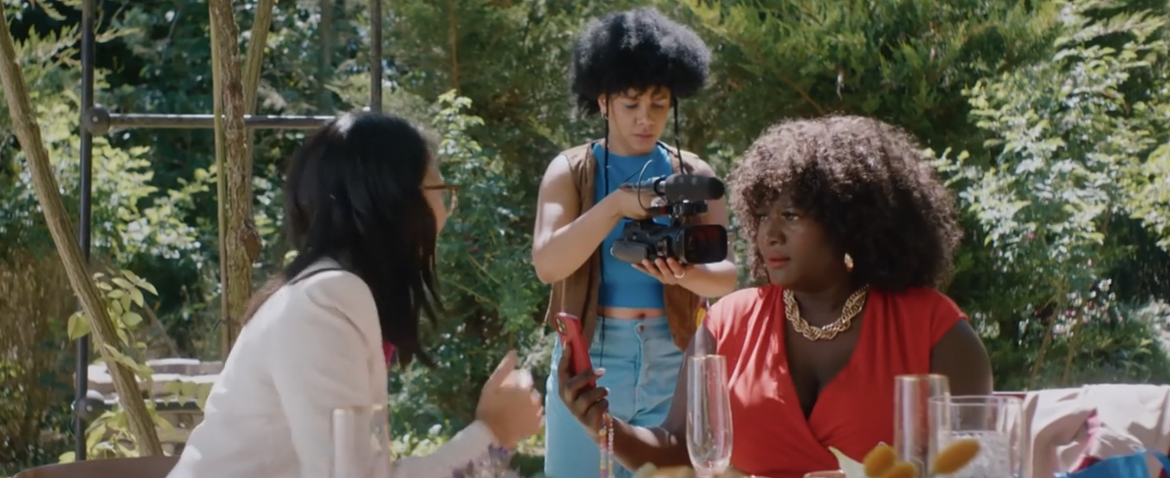 Zwei Personen sprechen am Tisch voll mit Gläsern. Eine im roten Kleid mit kinnlangen Locken und einem Handy. Eine andere von hinten mit Schwarzen glatten längeren Haaren. Im Hintergrund eine Person mit brauner Weste und kurzem Afro, arbeitet konzentriert mit einer Kamera.