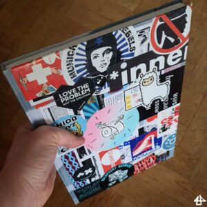 Einband des Skizzenbuches mit der linken Hand gehalten, es ist über und über mit aktivistischen Aufklebern beklebt, zum Beispiel gegen Burschenschaften oder für trans Rechte.