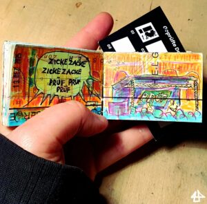 Foto eines winzigen Skizzenbuches in einer Hand, oben guckt die Stempelkarte der Prüf Demo raus. Das Bild mit Neonhighlightern, Buntstiften und schwarzem Fineliner. Eine Menschenmenge vor der Bühne mit zwei Sprechenden an, rechts über den Köpfen eine Neogelbe Sprechblase: Zickezacke, zickezacke, prüf, prüf, prüf.