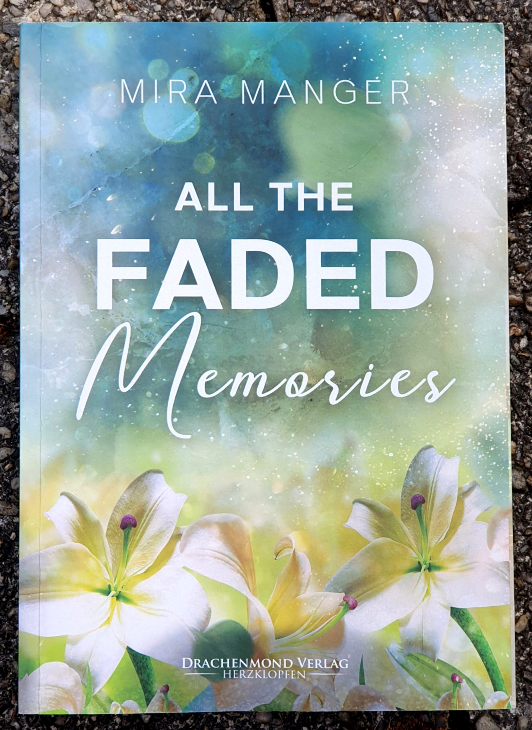 Foto des Buchdeckels von All the faded Memories von Mira Manger, Drachenmond Verlag Herzklopfen. Die Schrift ist weiß über die ganze Seite verteilt, im Hintergrund blaue und weiße Bereiche wie aquarelliert und unten drei weiße Lilien.