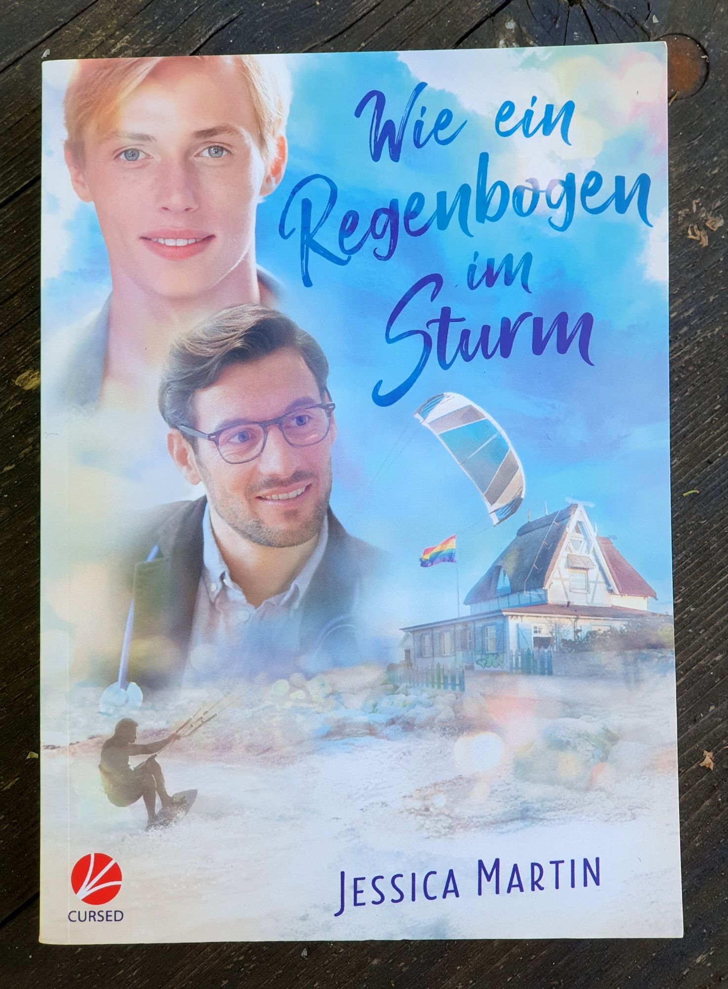 Buchdeckel von Wie ein Regenbogen im Sturm von Jessica Martin im Cursed Verlag. Ein zwanzigjähriger Blonder, ein dreissigjähriger mit runder Brille und der Schattenriss eines Kite Surfers vor einem kleinen Strandhotel an dem eine Regenbogenflagge weht. Alles realistisch in Pastelfarben gezeichnet.